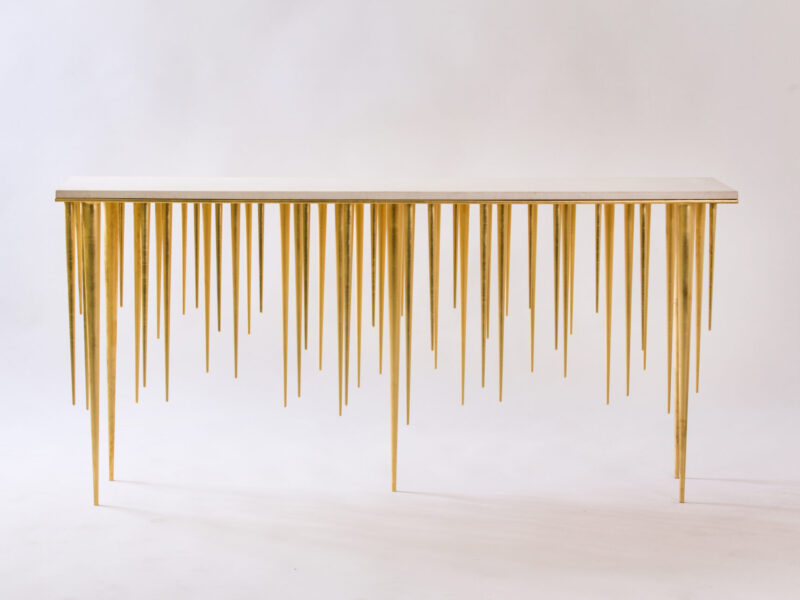 Golden console table with many slender, pointed legs resembling spikes against a plain white backdrop, minimalist gallery style.