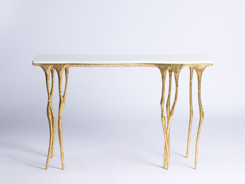 White rectangular tabletop with ornate gold sculpted legs set against a soft light gradient background, minimal styling piece leant against a wall.