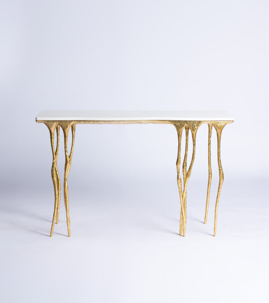 White rectangular tabletop with ornate gold sculpted legs set against a soft light gradient background, minimal styling piece leant against a wall.