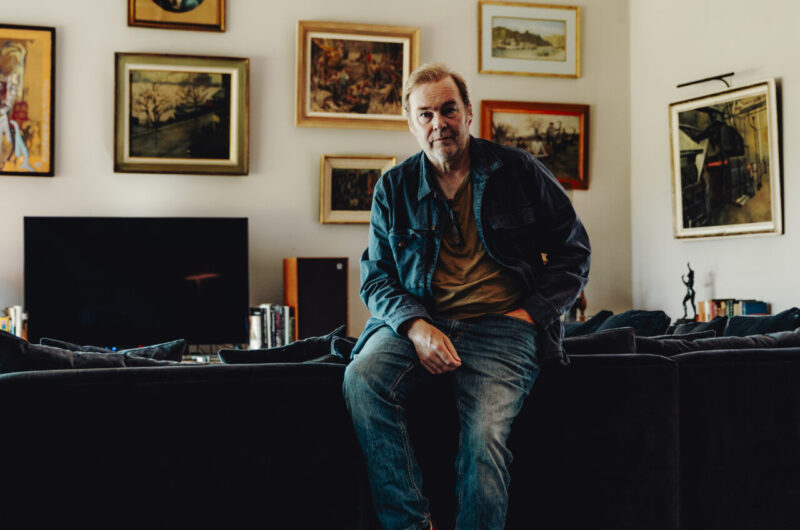 Man sitting on a dark couch in a living room, wall full of framed artworks, two dogs resting on a fluffy rug in the foreground.