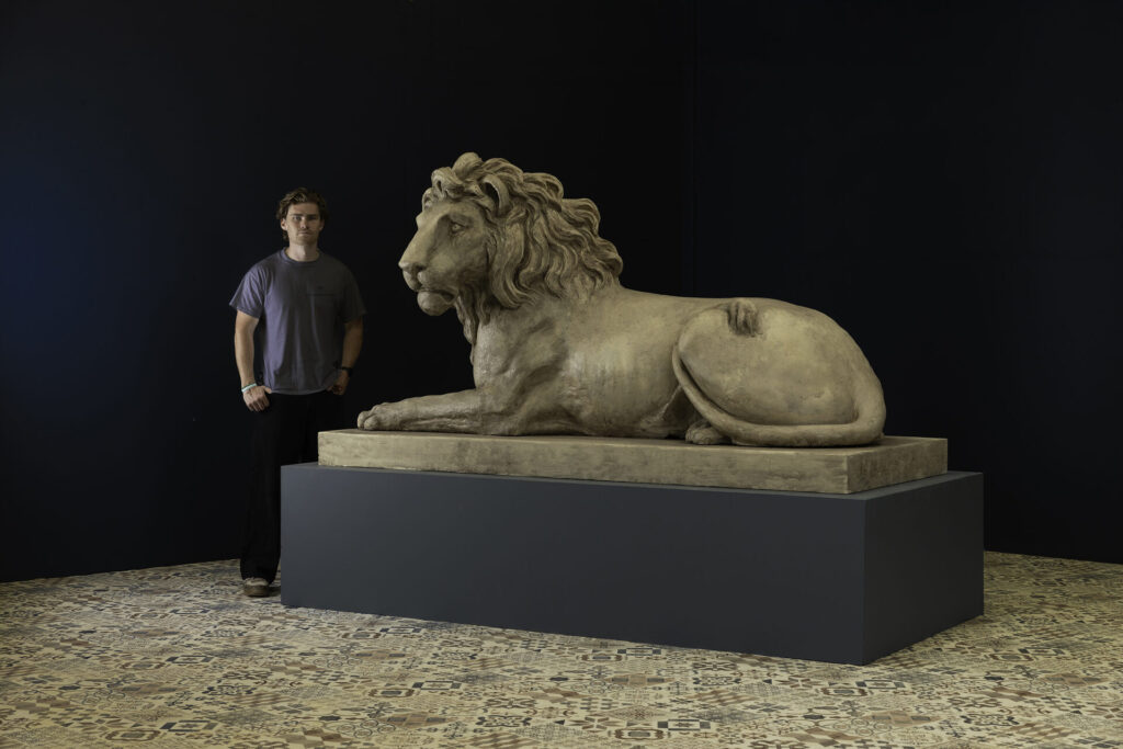 Man standing next to a large reclining lion sculpture on a pedestal in a museum, dark walls behind.