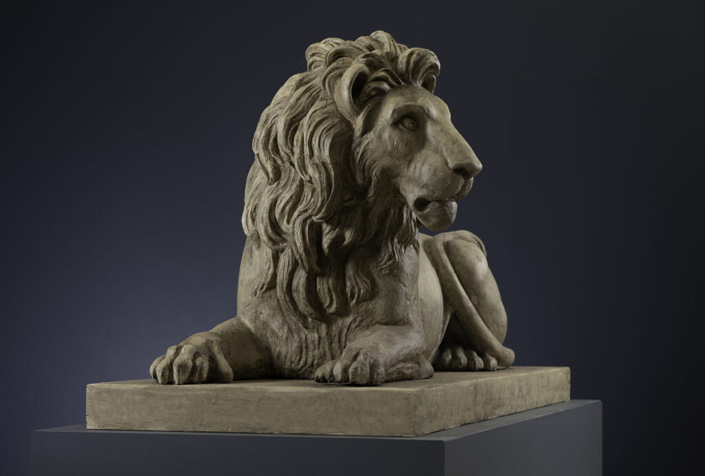 Stone sculpture of a reclining lion with front paws on a pedestal against a dark background.