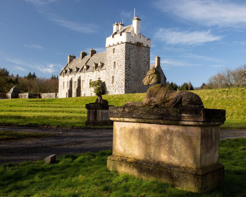 Stone castle-like building with crenellated tower on a grassy field; a Stone Kingsale hound dog statue sits in the foreground.