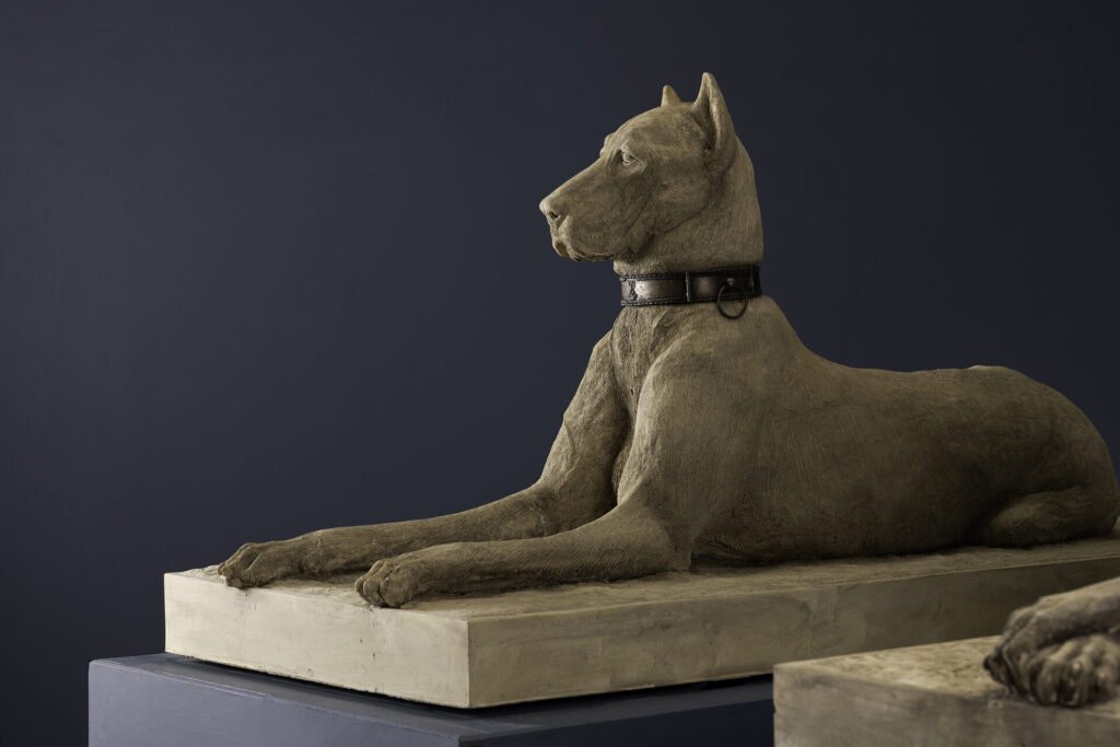 Lying dog statue with a leather collar on a stone pedestal against a dark wall, art sculpture view.