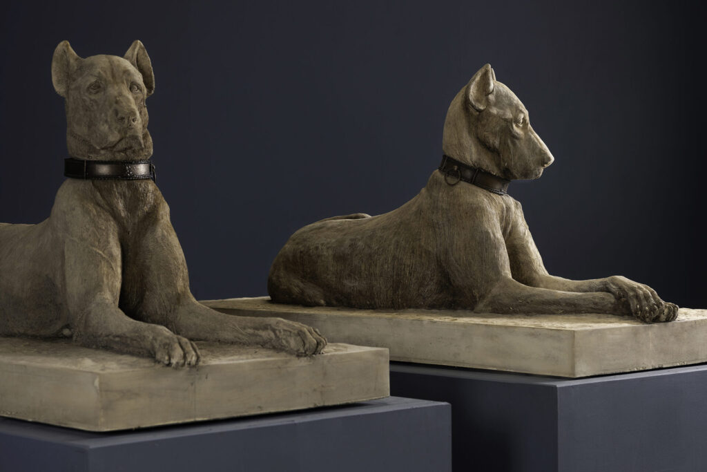 Two stone dog statues with collars resting on a light pedestal against a dark background, facing slightly apart.