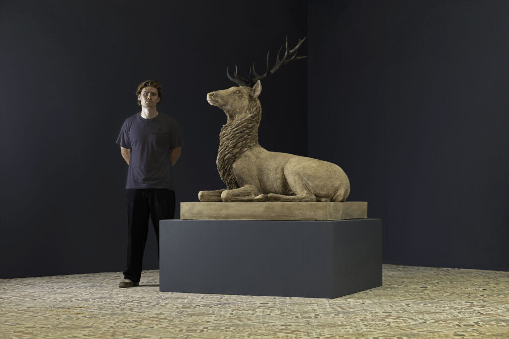 A man stands beside a large, reclining deer statue with antlers on a pedestal in a gallery-like room with dark walls.
