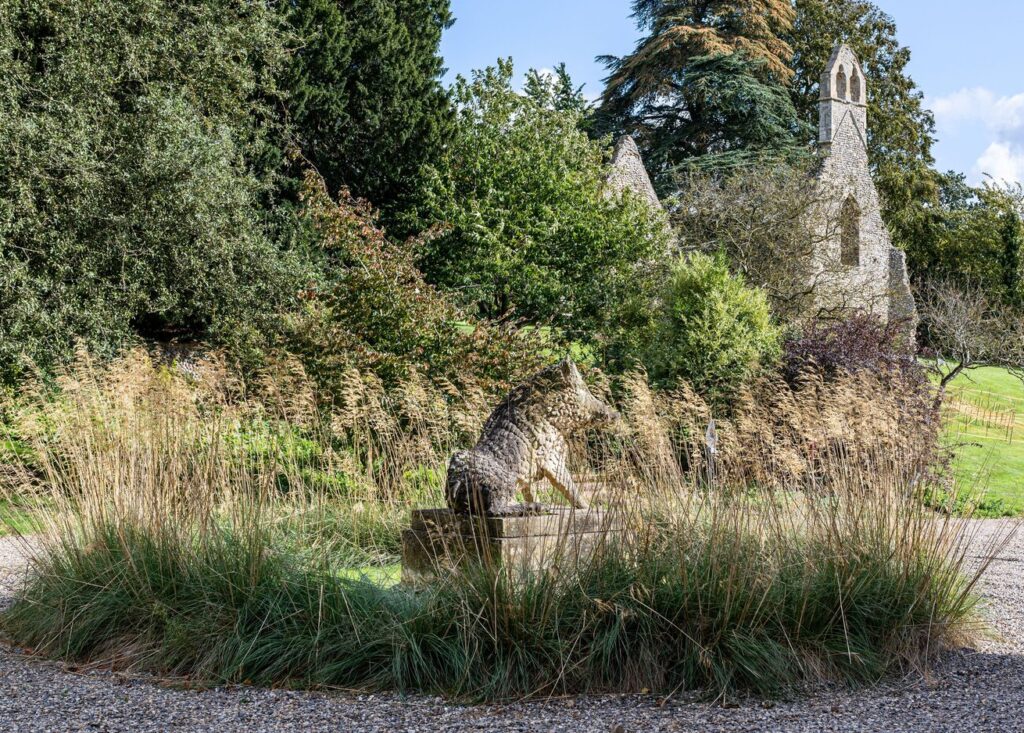 Stone uffizi boar garden statue by a Ruined stone church with small bell tower among trees, viewed from a gravel circle with tall ornamental grasses in a park-like setting.