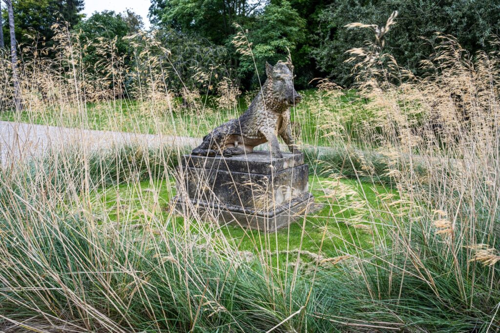Stone uffizi boar garden statue on a pedestal in a garden, framed by tall beige ornamental grasses.