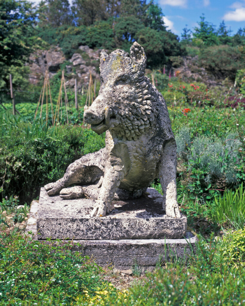 Stone uffizi boar garden statue perched on a weathered pedestal in a lush garden with greenery and flowers in the background.