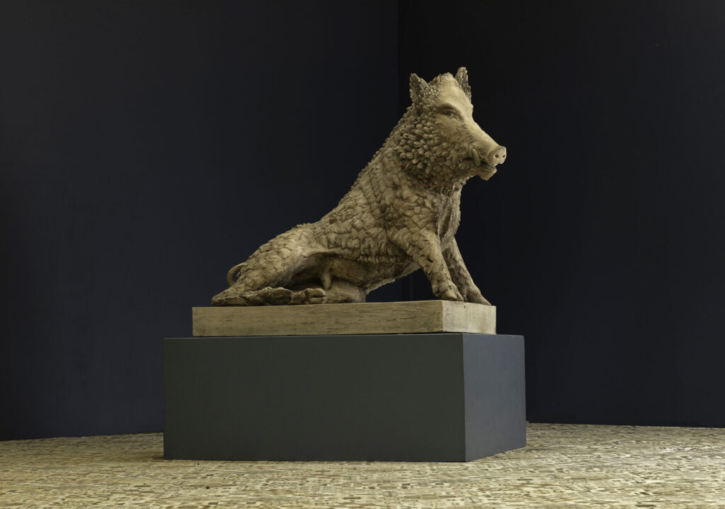 Stone uffizi boar sculpture seated on a pedestal in a dim gallery with a dark wall backdrop.