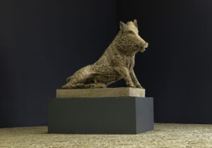 Stone uffizi boar sculpture seated on a pedestal in a dim gallery with a dark wall backdrop.