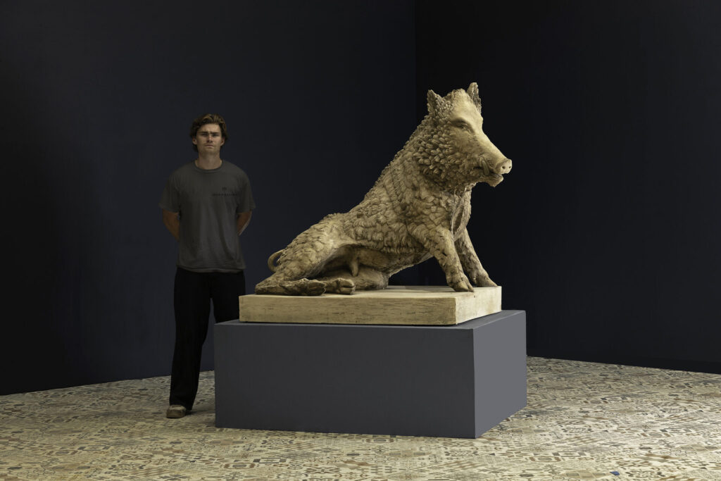 Young man in a gray shirt stands beside a large stone wolf sculpture on a pedestal in a gallery setting.